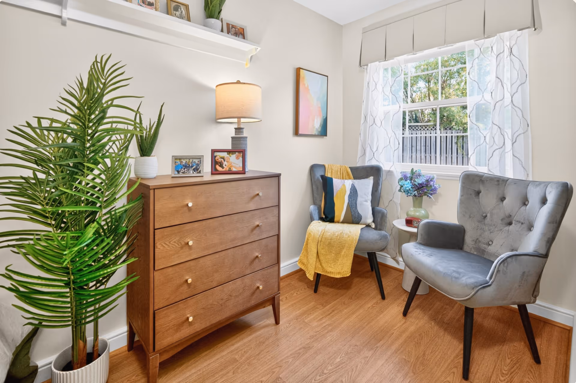 Cozy living room corner with two chairs, a dresser, plants, and a sunlit window.