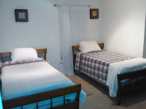 Two twin beds in a simple, sparsely decorated bedroom with a sheer curtain and framed pictures on the wall.