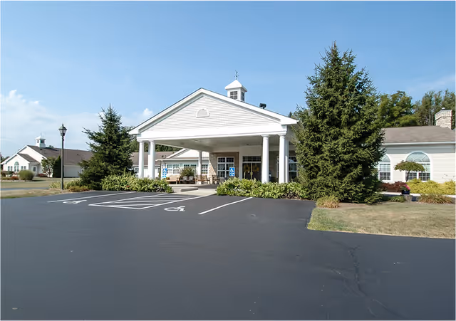 Front exterior view of a single-story senior living facility building with a covered entrance supported by white columns, surrounded by greenery and a paved parking area with handicap parking spaces.