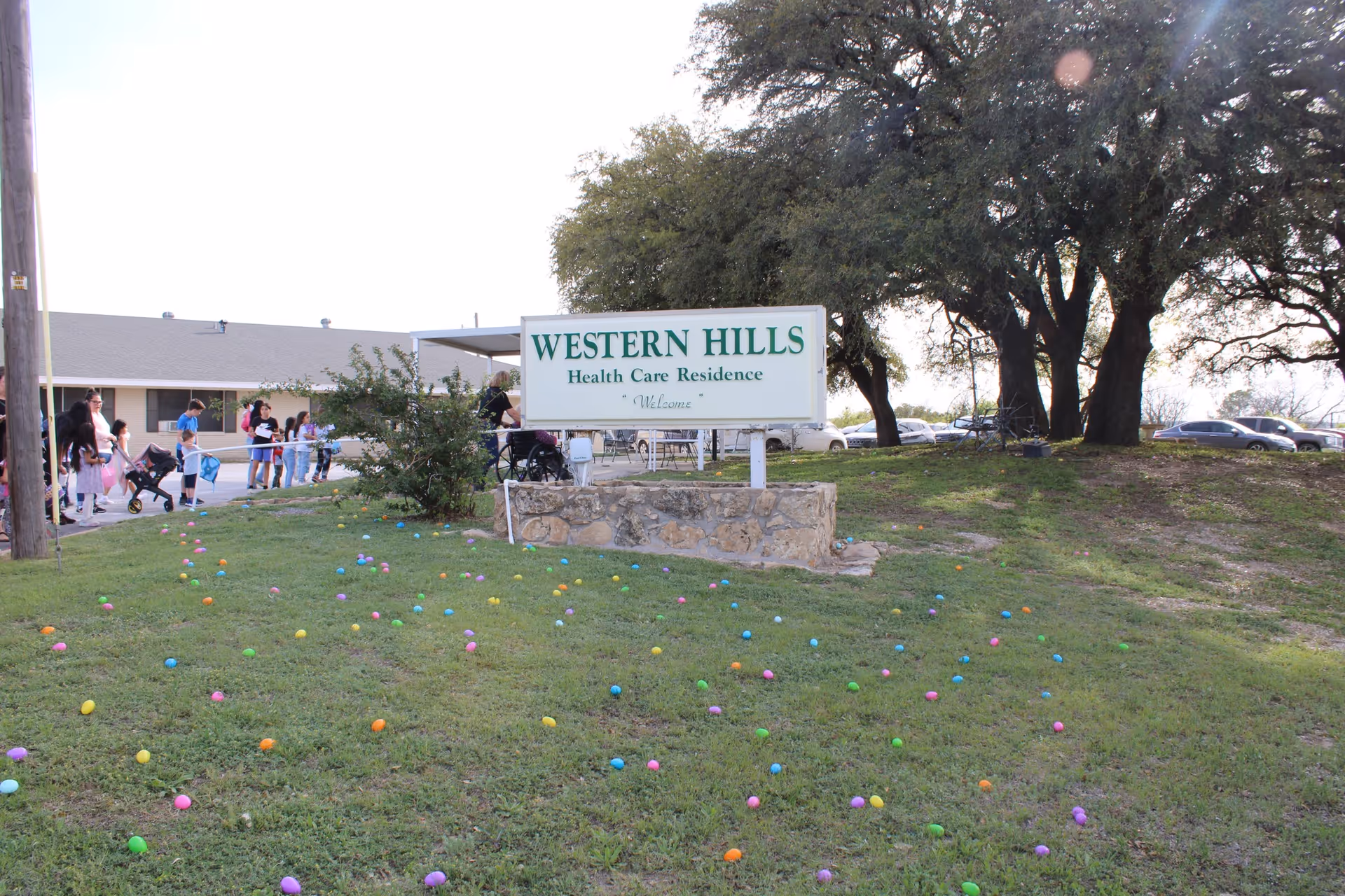 Outdoor view of Western Hills Health Care Residence with a sign in the foreground on a grassy area scattered with colorful plastic Easter eggs. In the background, there are people, including children and adults, near the building and trees.