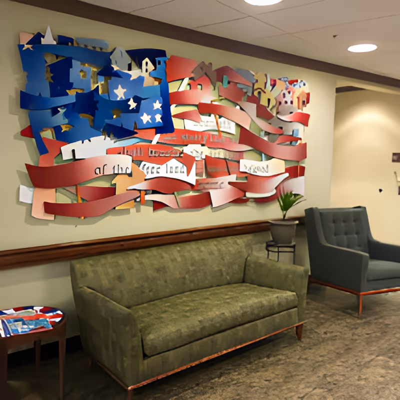 A seating area in a facility with a green couch and a gray armchair. Above the couch is a large decorative wall art piece resembling an abstract American flag with red, white, and blue colors. A small side table with a colorful top and a potted plant on a stand are also visible. The walls are beige and the ceiling has recessed lighting.