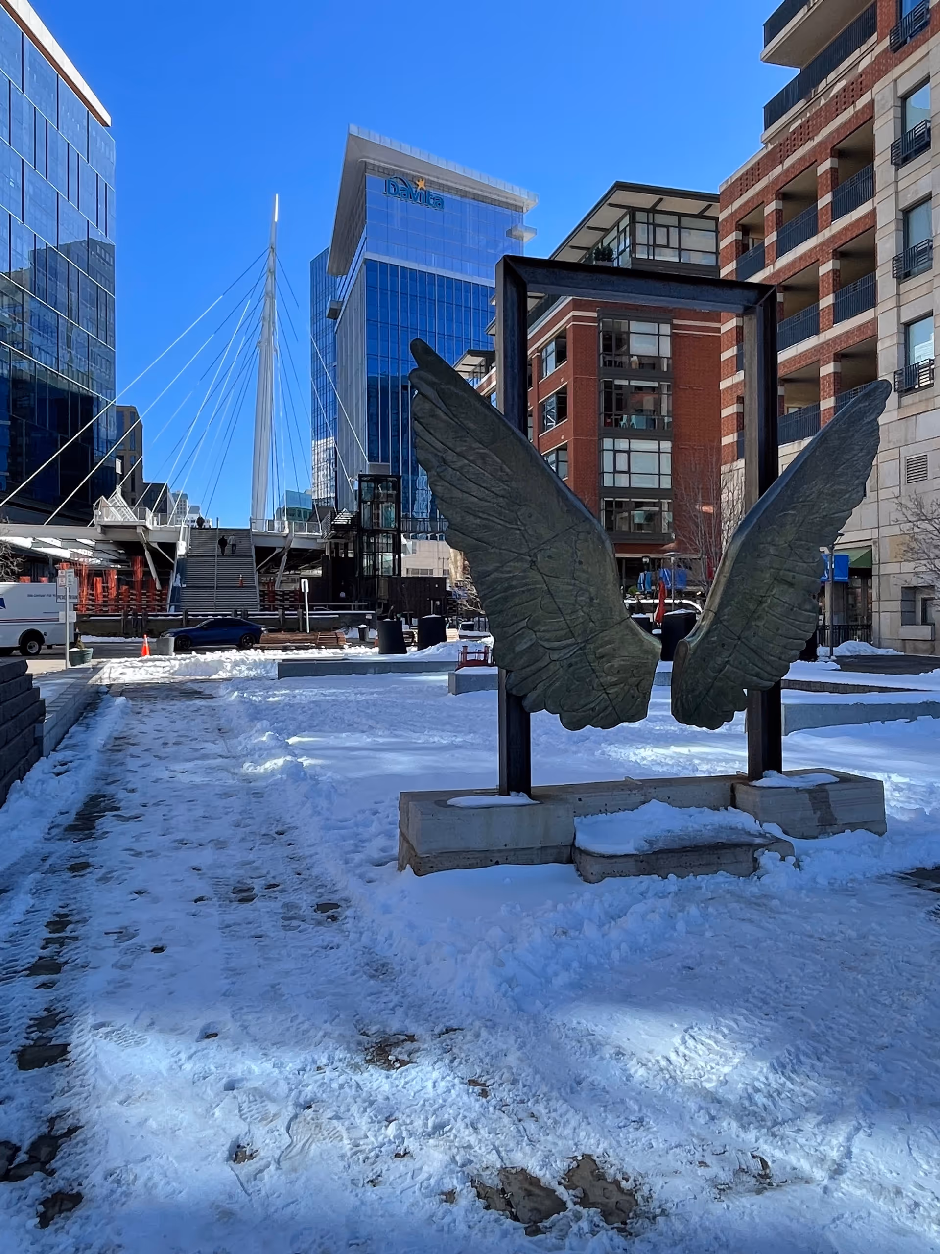 Outdoor plaza with a wing sculpture, modern glass buildings, and snow-covered ground.