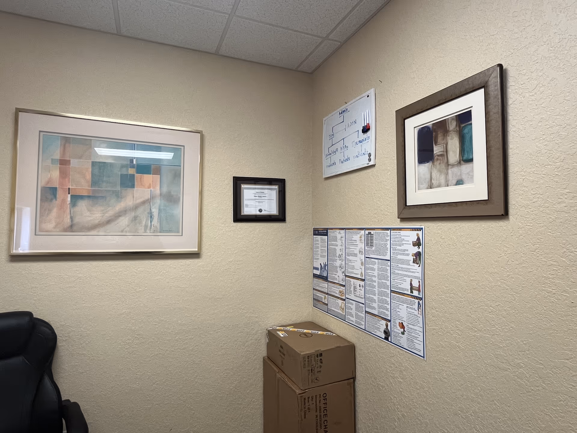 Corner of beige-walled office area with framed art, charts, and stacked boxes.