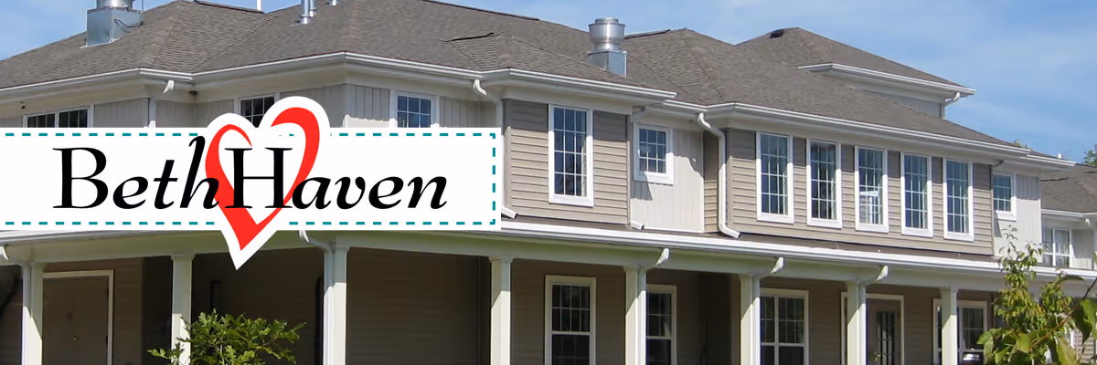Two-story beige assisted living building with many windows and a covered porch supported by white columns.