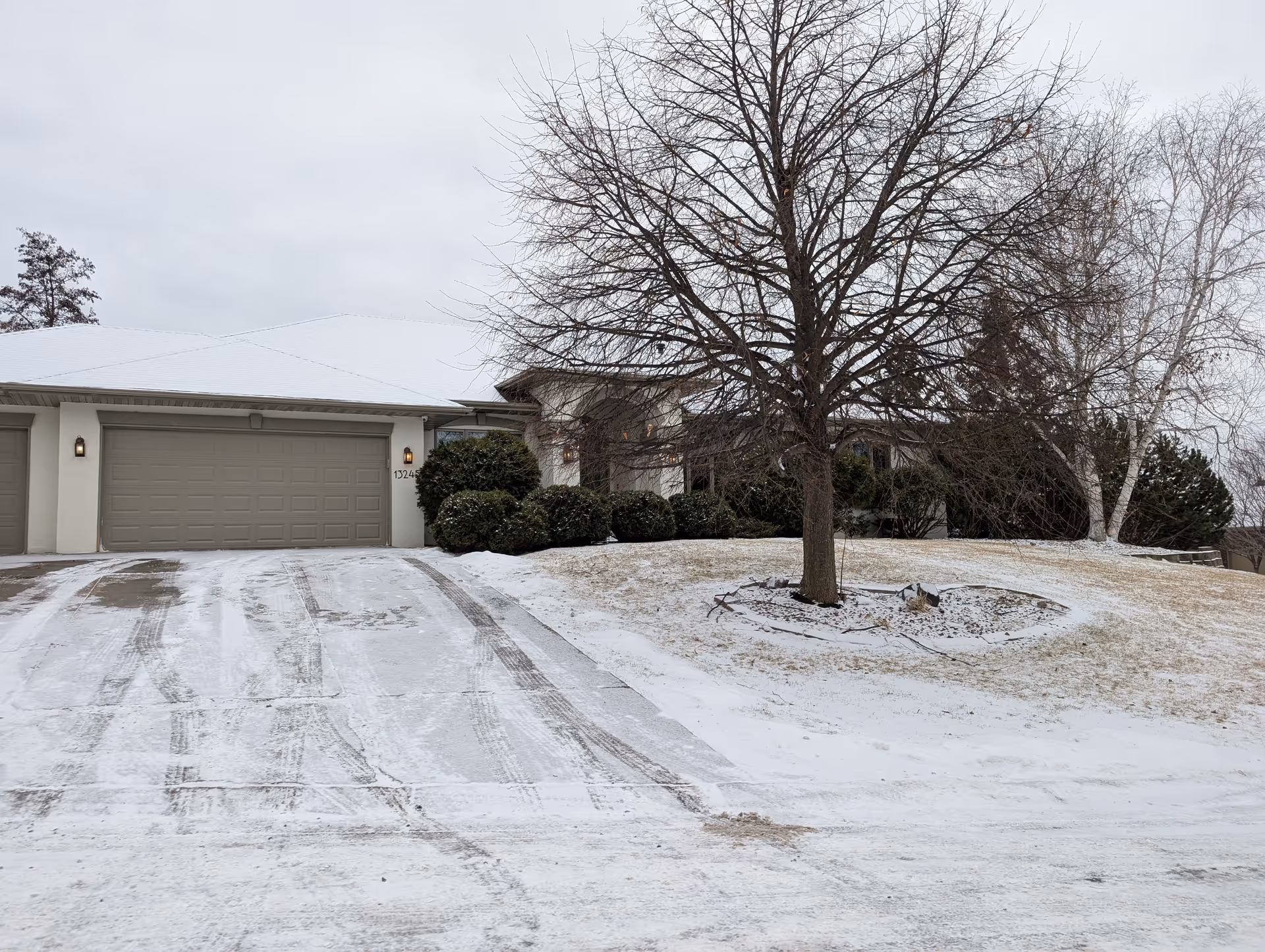 Front of a single-story residence with a three-car garage, snow-covered driveway, and a large leafless tree on the lawn.