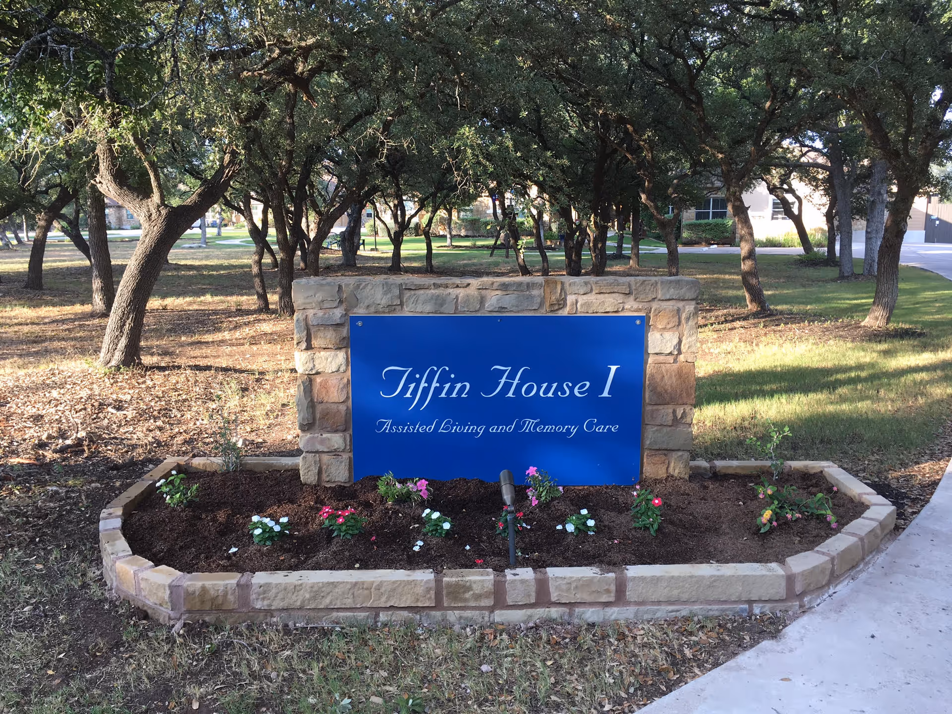 Stone-bordered flower bed with a blue Tiffin House sign in a tree-lined front area.