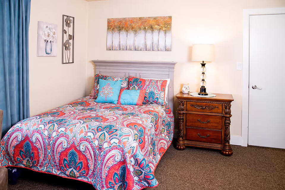A bright bedroom with a colorful paisley bedspread, decorative pillows, a wooden nightstand with a lamp, and wall art above the headboard.