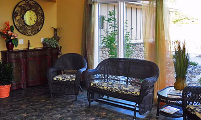 Cozy seating area with wicker chairs and a loveseat, a side table, decorative cabinet and a large window letting in natural light.