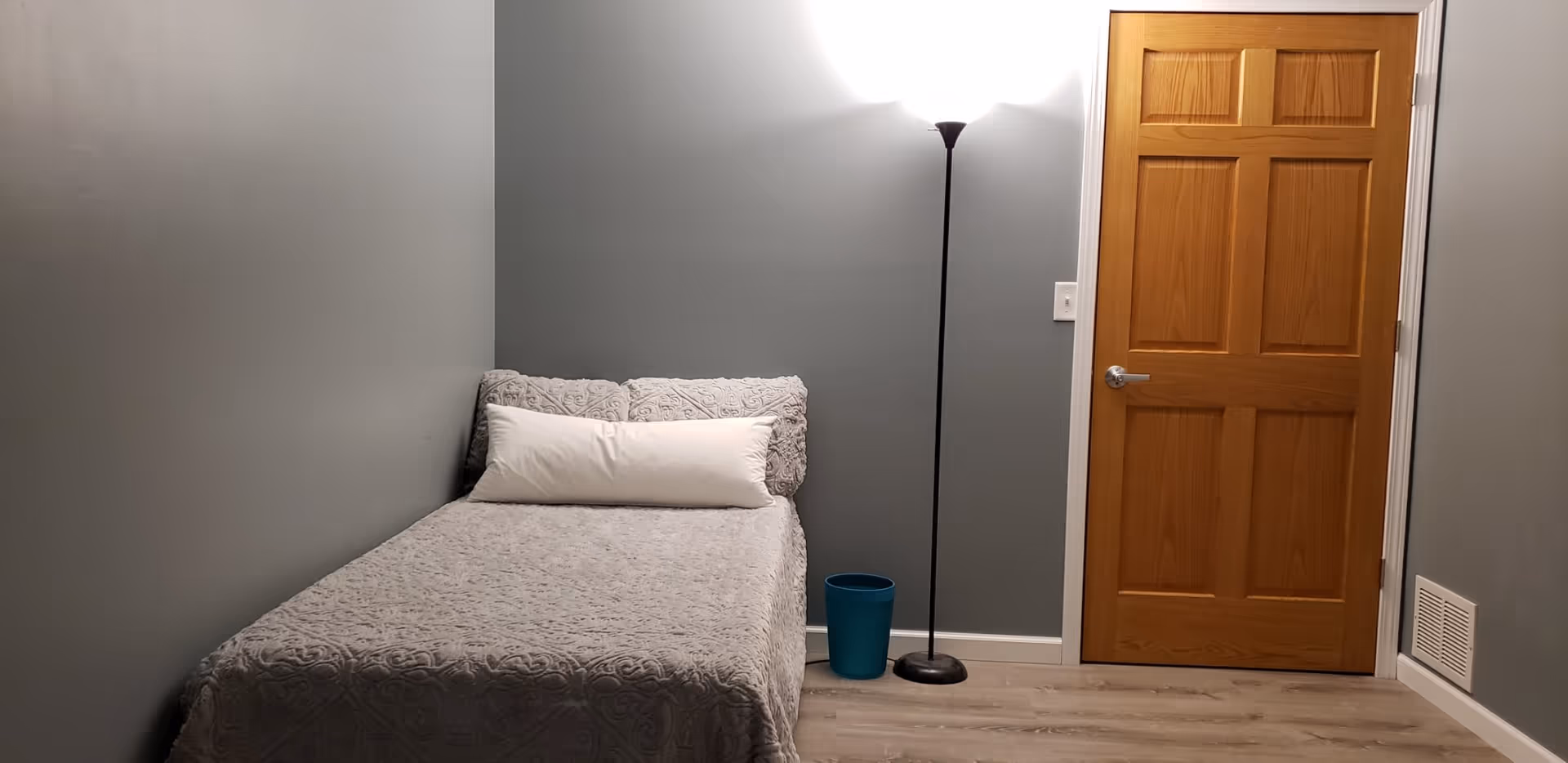 A small bedroom with gray walls and light wood flooring. There is a single bed with a gray patterned bedspread and a long white pillow. Next to the bed is a tall black floor lamp emitting bright light and a blue waste bin. A wooden door with six panels is on the right side of the room.