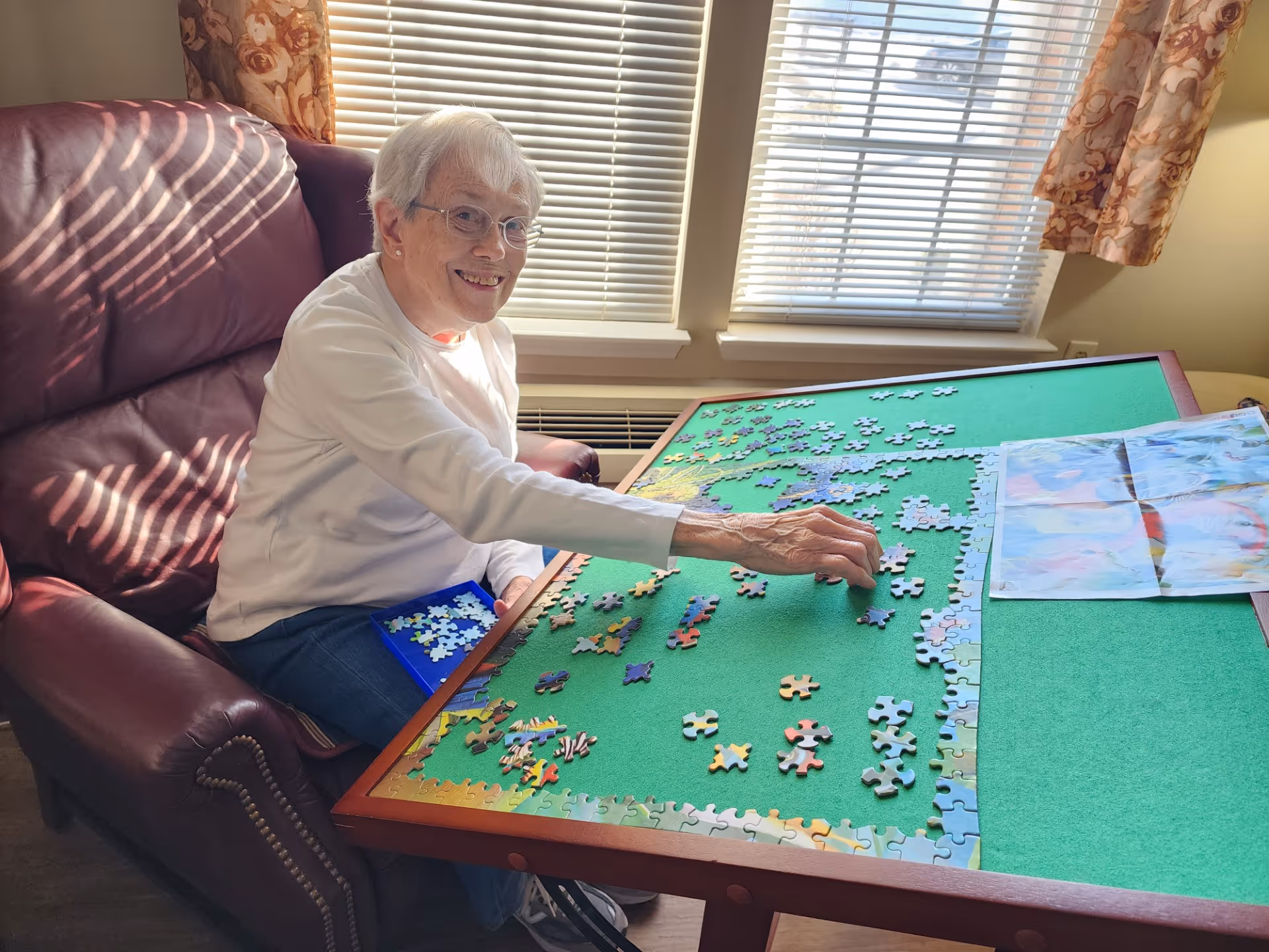 An elderly woman with short white hair and glasses is sitting on a maroon leather armchair next to a large green puzzle table. She is smiling and placing a puzzle piece on the table, which has many scattered puzzle pieces and a partially completed border. Behind her are two windows with blinds and floral curtains.