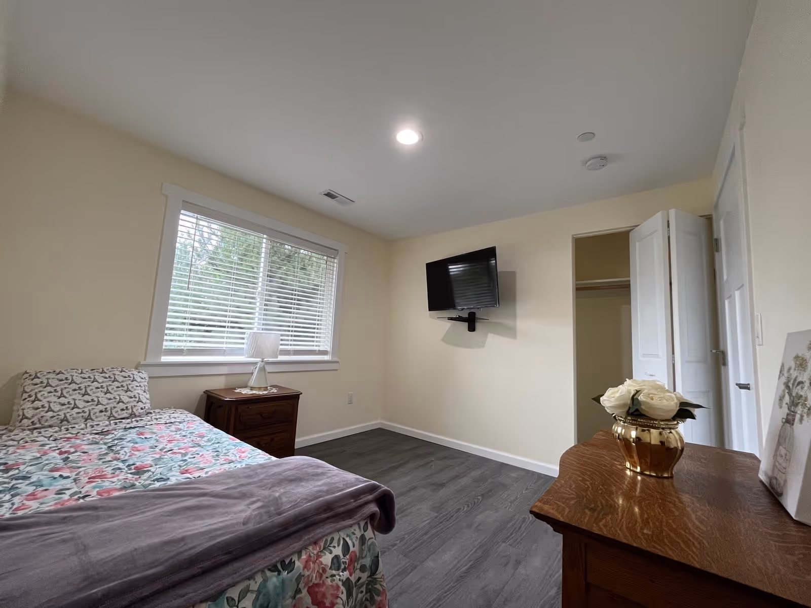 A furnished bedroom with bed, nightstand, dresser, window, and wall mounted TV in a senior facility.