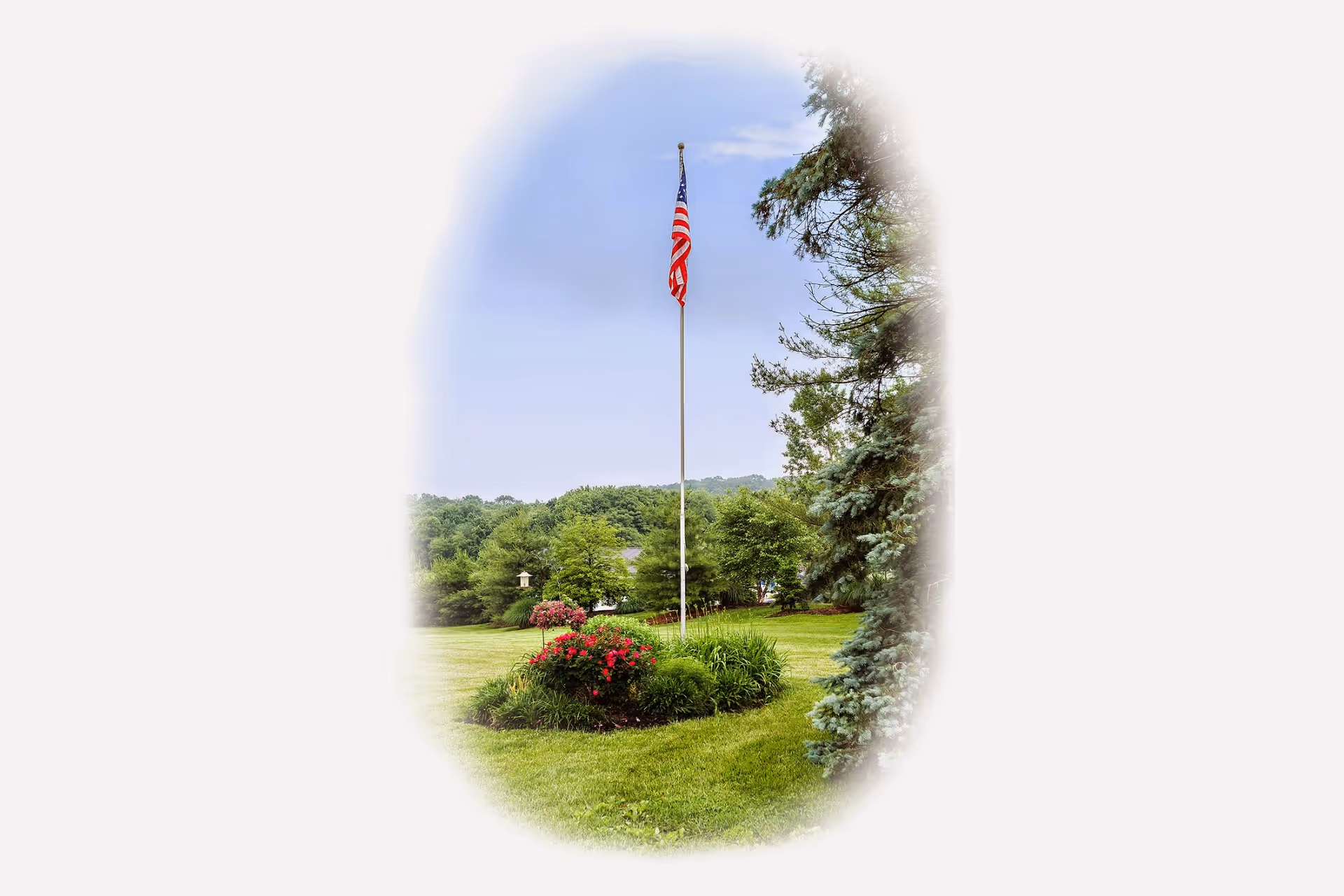 A tall flagpole with an American flag flying at the top stands in the center of a well-maintained grassy area surrounded by flowering bushes and trees, with a clear sky and distant hills in the background.
