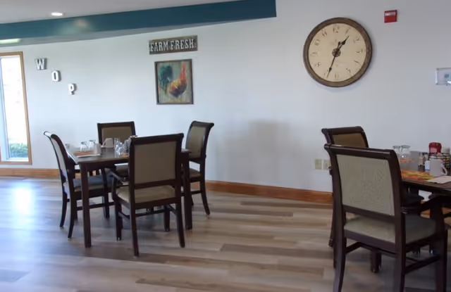 A dining room with tables set for meals, wooden chairs, and a large wall clock.