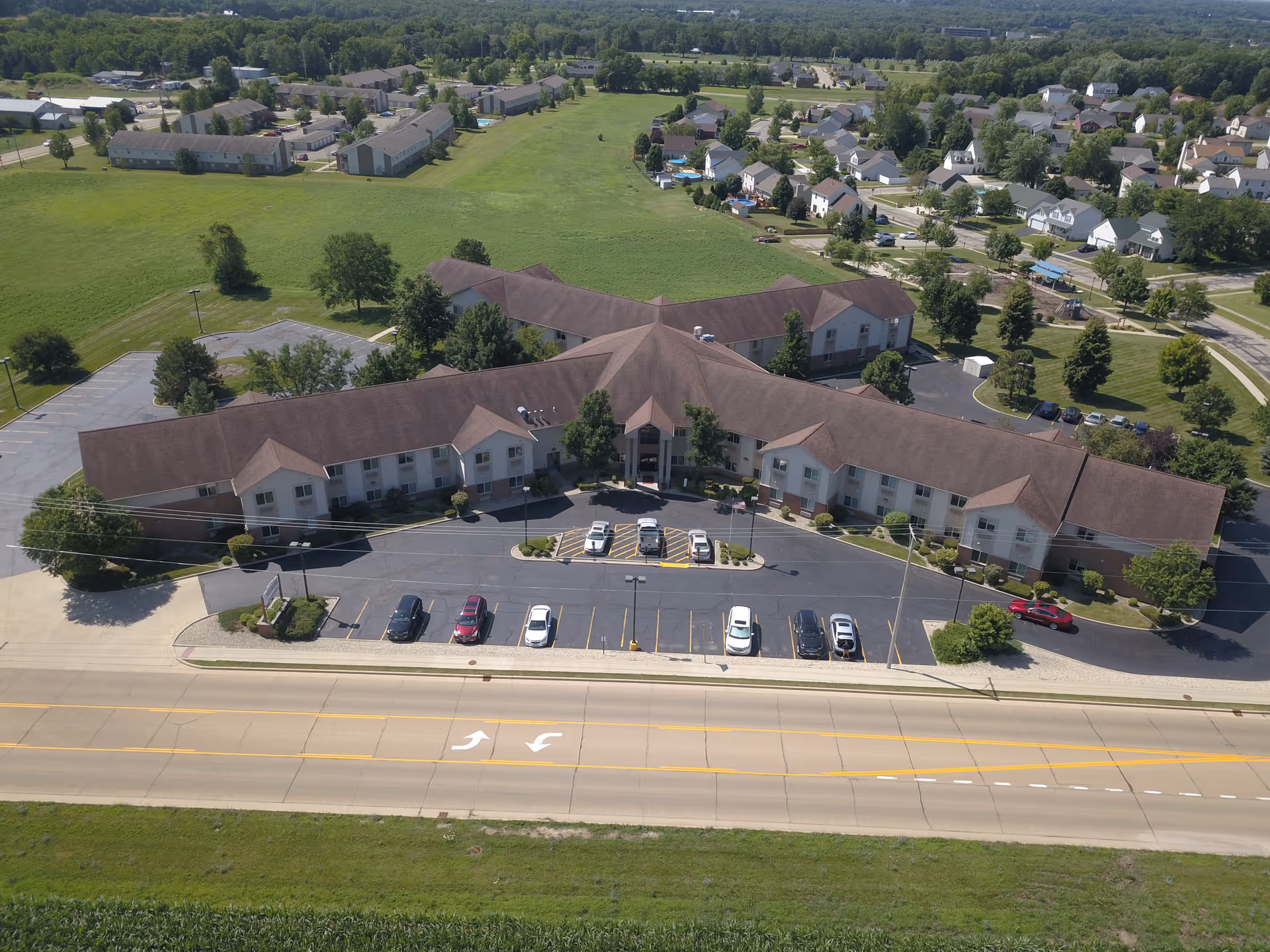 Aerial view of a large senior living facility building with a brown roof and light-colored walls, surrounded by parking lots with several cars parked. The building is situated in a green area with trees and grass, and residential houses are visible in the background.