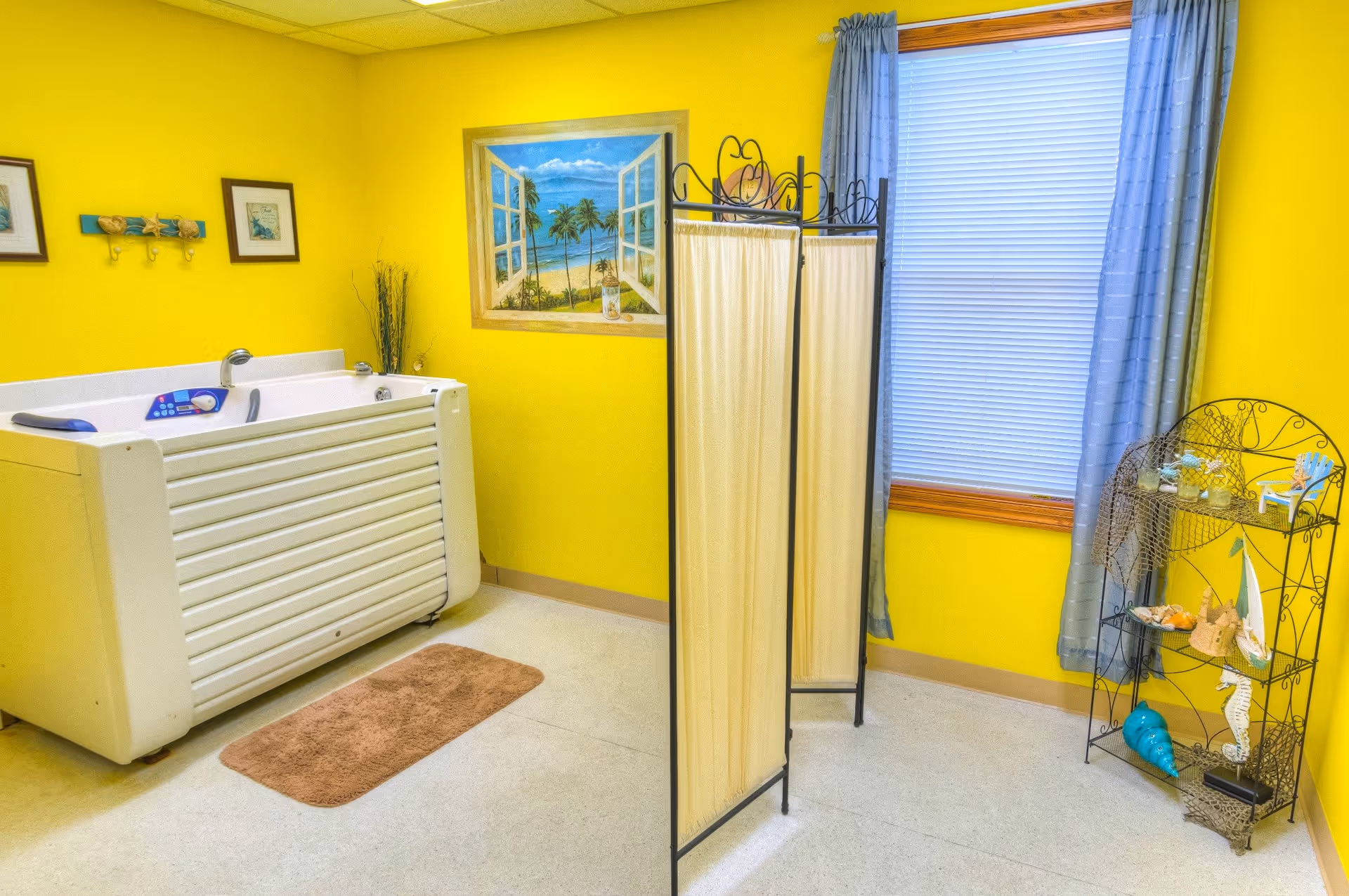 A bright yellow room with a white walk-in bathtub, a brown bath mat, a folding privacy screen, a window with blue curtains, and a decorative metal shelf holding seashell and beach-themed items. There are also framed pictures and a wall painting of a beach scene.