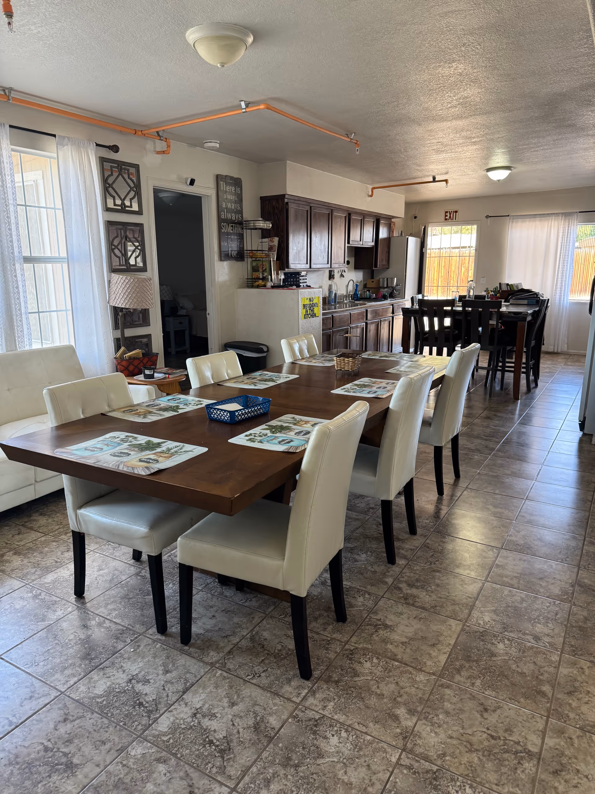 Interior view of a dining area in Rose Garden Assisted Living featuring a large wooden dining table with eight white cushioned chairs. The table is set with placemats and a small basket. In the background, there is a kitchen area with dark wooden cabinets, a refrigerator, and a smaller dining table with chairs near a window with white curtains. The floor is tiled, and the room is well-lit with natural light coming through the windows.