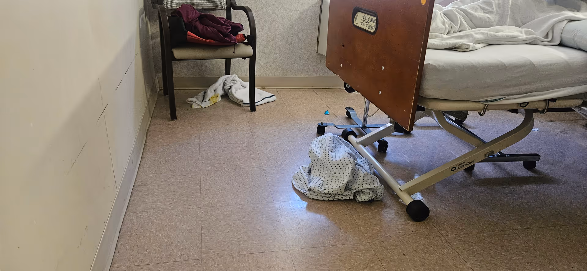 Hospital-style patient room with a bed, chair, and scattered clothing on linoleum floor.