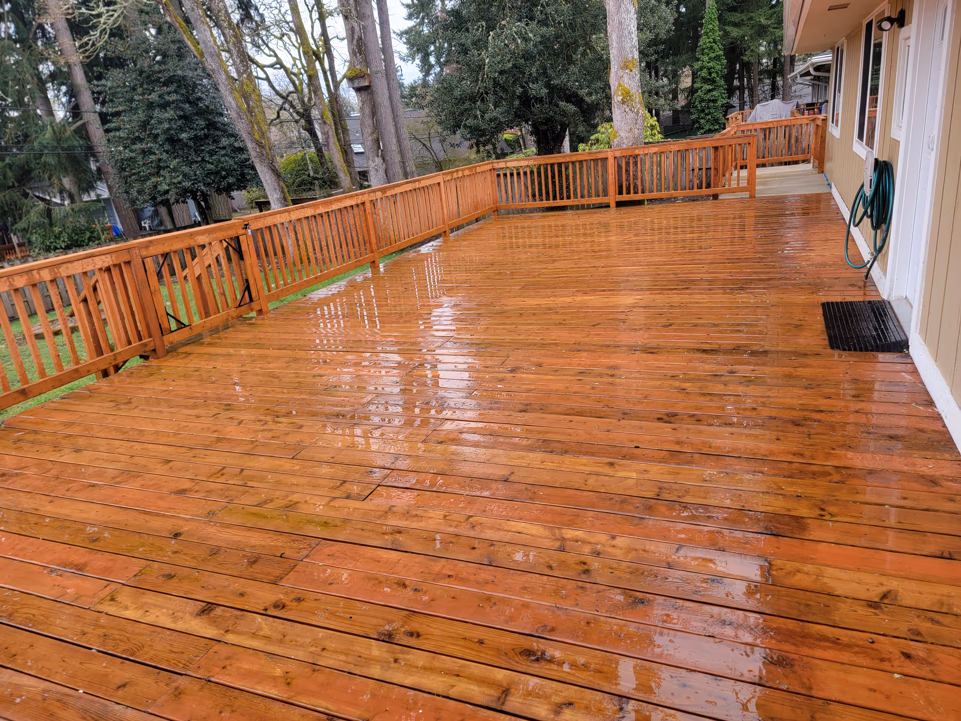 Rain-soaked wooden deck beside a beige home, with railing and trees in the background.