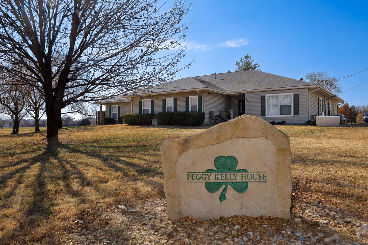 A single-story building with beige siding and green shutters, surrounded by a grassy area with leafless trees. In the foreground, there is a large rock with a green shamrock and the text 'PEGGY KELLY HOUSE' engraved on it. The sky is clear and blue.