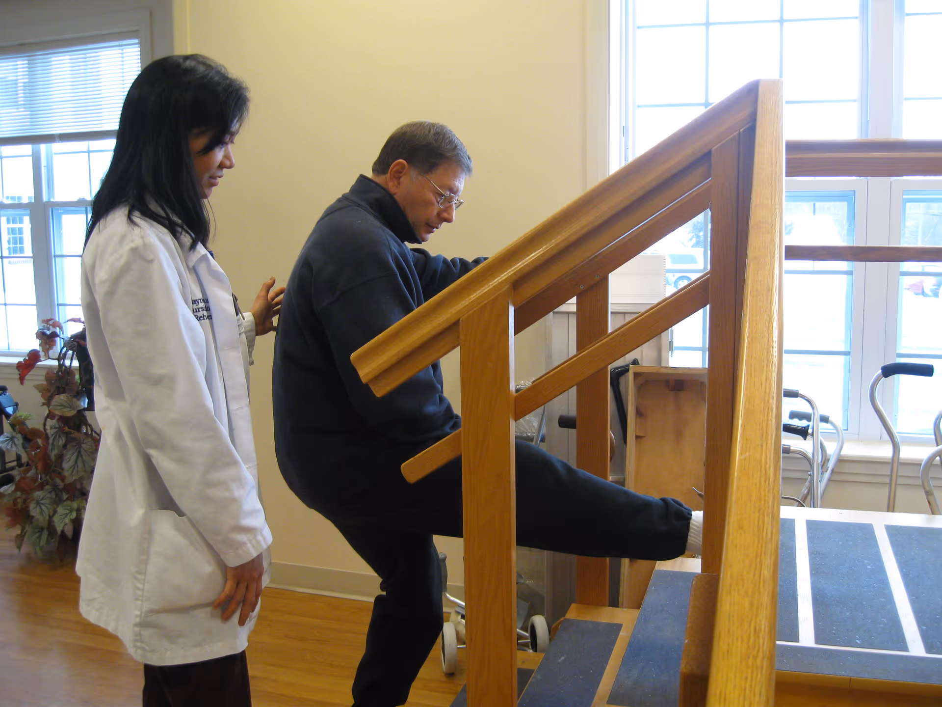 A healthcare professional in a white coat assists an elderly man who is exercising by stepping onto a wooden staircase with handrails inside a well-lit room with large windows.