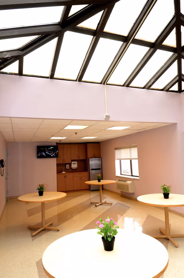 Bright dining area with round tables, potted plants, kitchenette, and skylit ceiling.