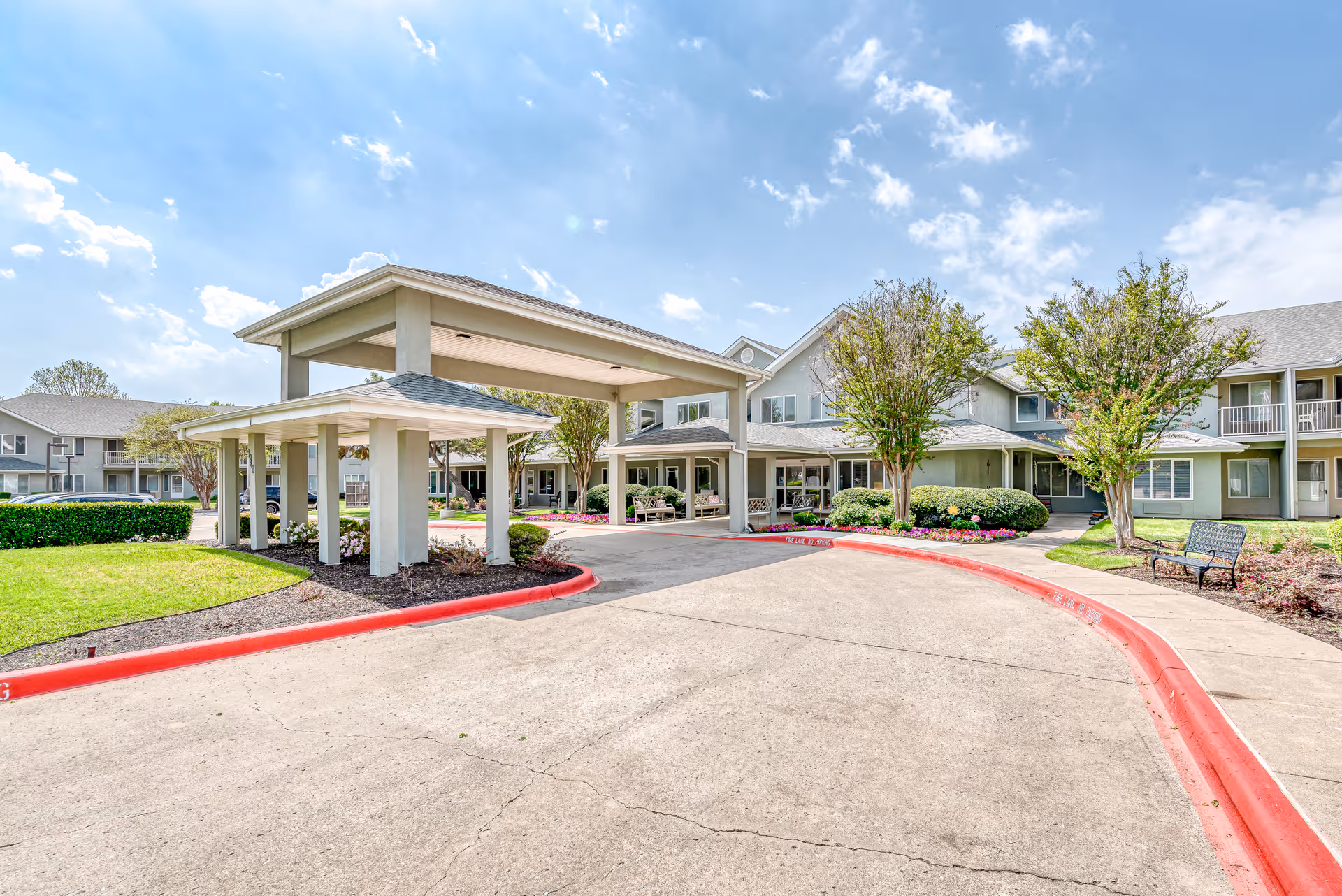 Exterior view of Solstice Senior Living at Plano showing a covered entrance with a driveway, landscaped greenery, trees, and a bench under a partly cloudy sky.