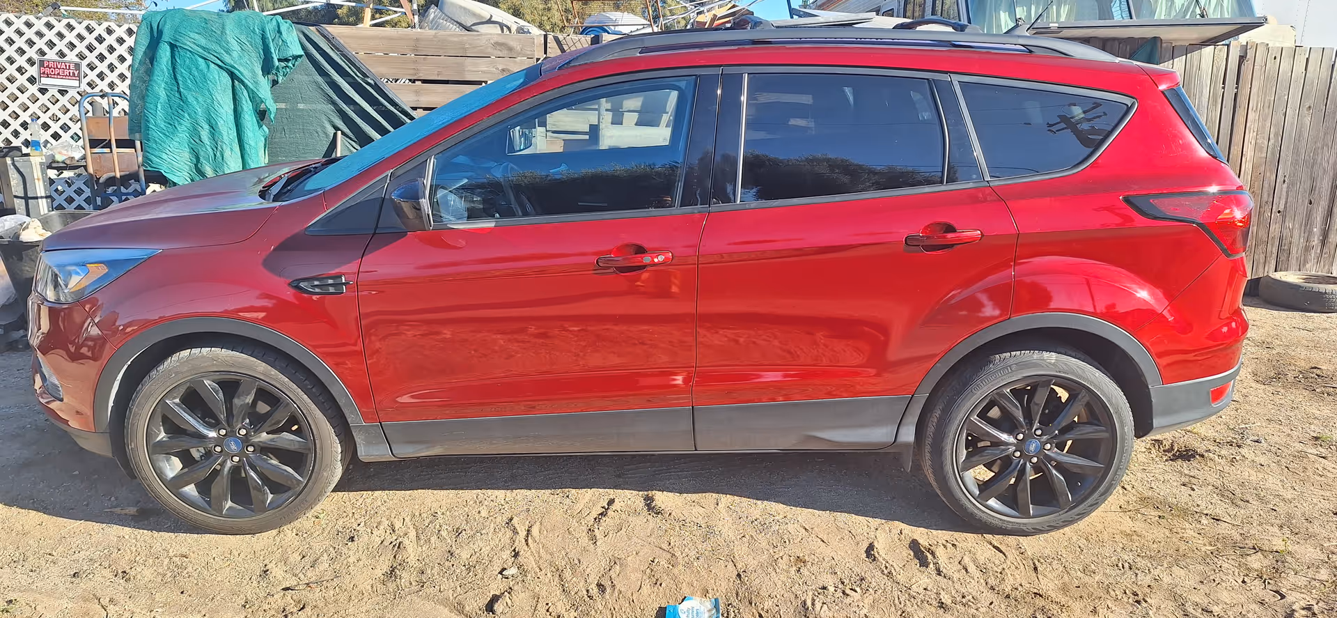 Red SUV parked on a dirt surface beside a wooden fence and scattered items.