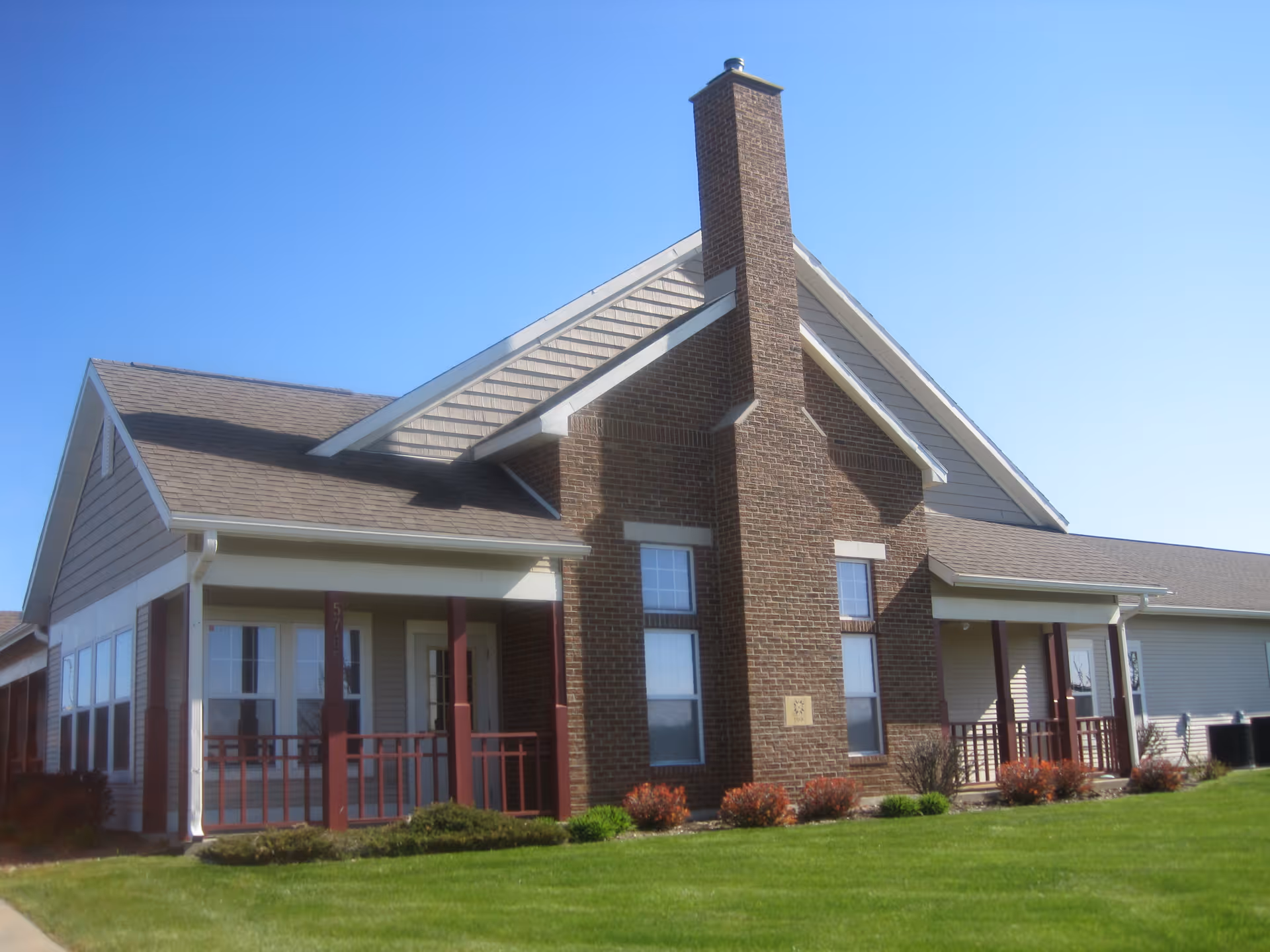 Exterior view of a single-story residential building with a brick chimney, beige siding, and a covered porch with red railings. The building is surrounded by a well-maintained green lawn and small shrubs under a clear blue sky.