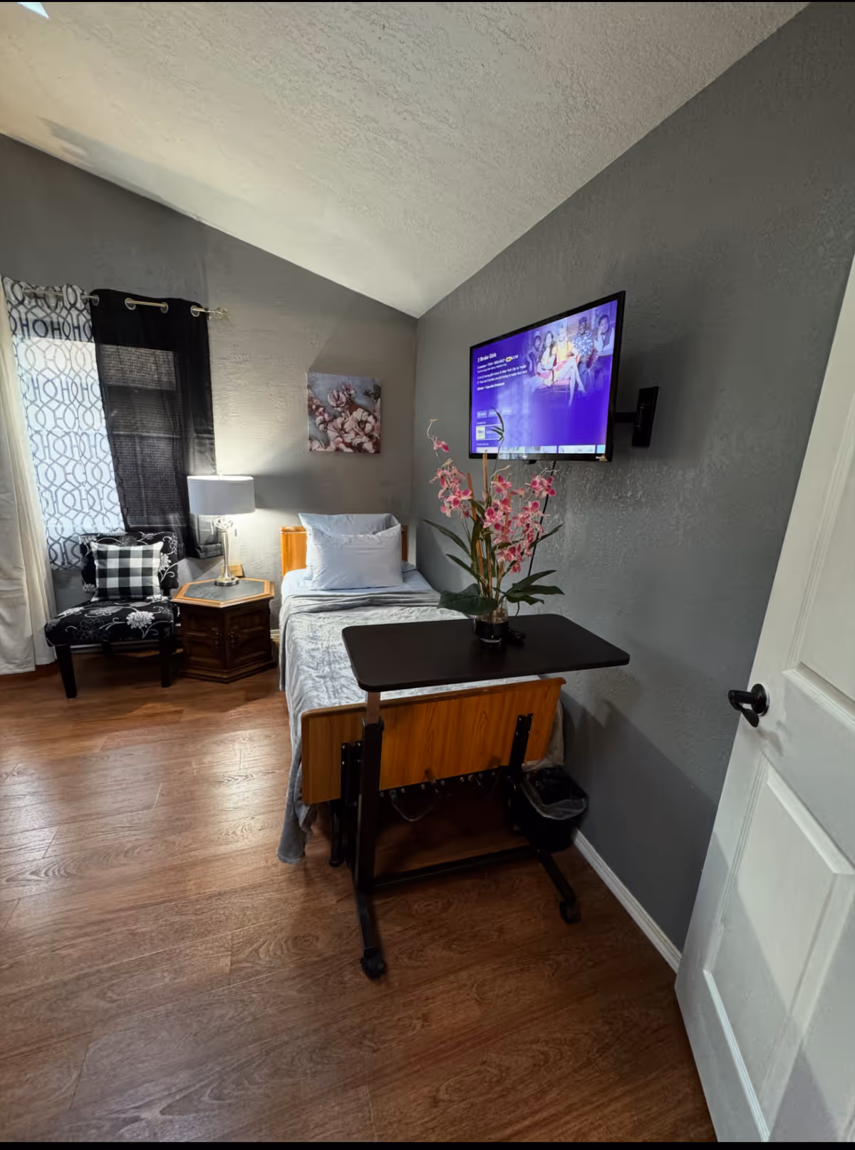 Small patient bedroom in a senior facility with bed, nightstand, desk, chair, and mounted TV.