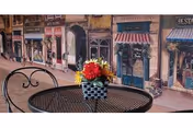 Round black metal outdoor patio table with a red-yellow flower centerpiece, storefront mural background.