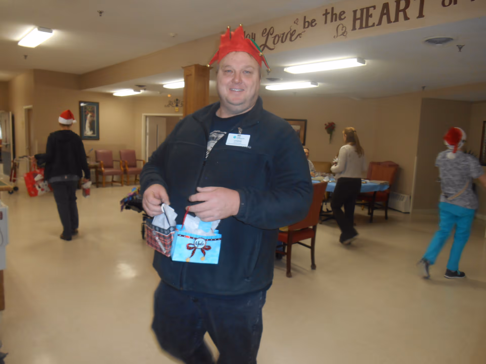 A man wearing a red festive hat and a black jacket is smiling and holding two small gift bags in a communal room. In the background, several people are walking or sitting around tables, some wearing Santa hats. The room has beige walls, chairs, and tables, with a decorative wall decal that reads 'Love be the HEART'.