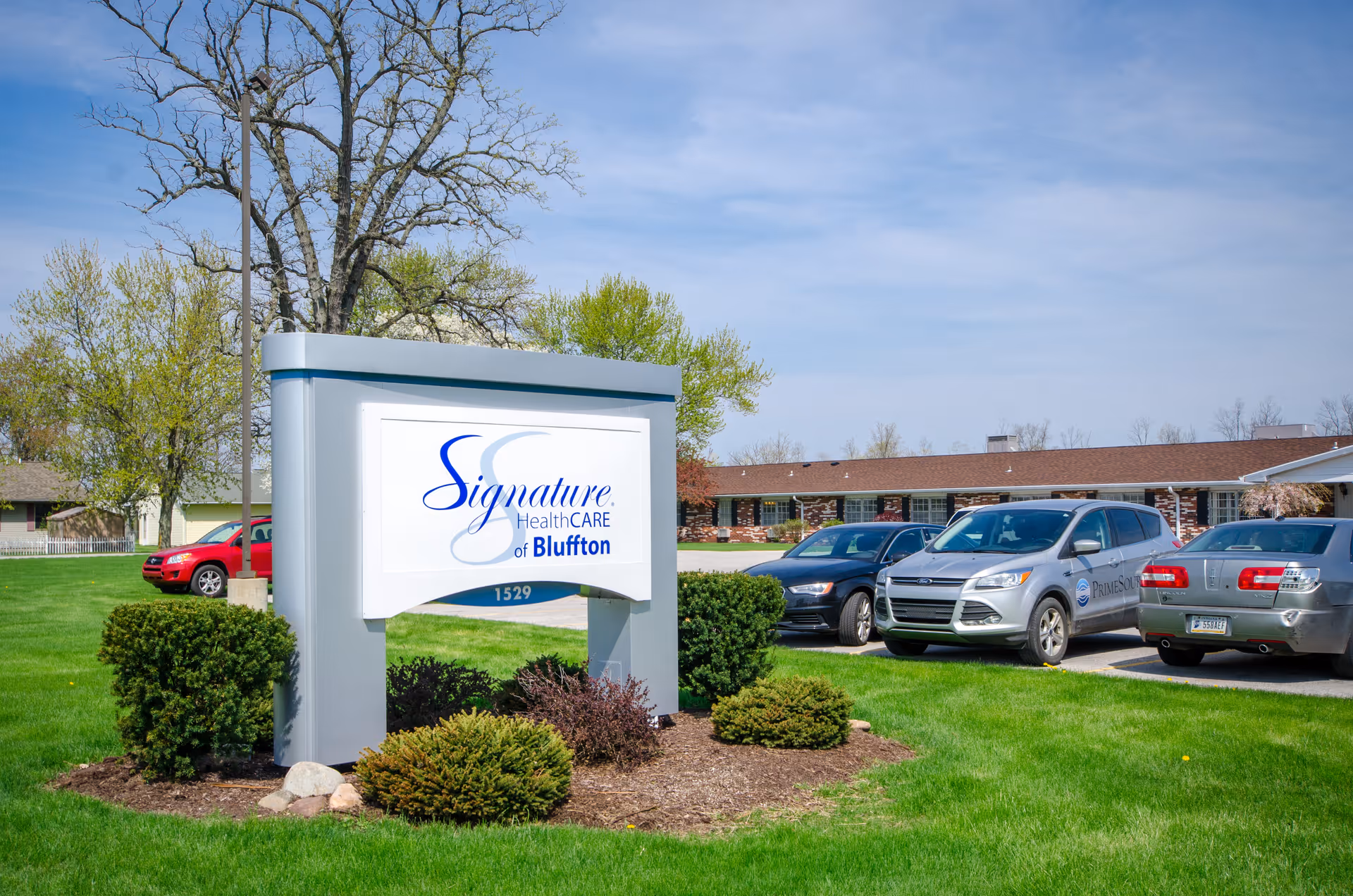 Outdoor view of the Signature HealthCARE of Bluffton facility sign with a parking lot and several cars in front of a single-story brick building, surrounded by green grass and trees under a blue sky.