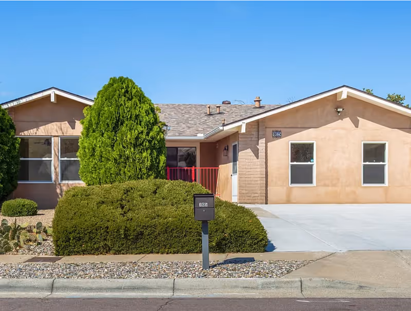 Single-story beige assisted living building with shrubs, a driveway, and a clear blue sky.