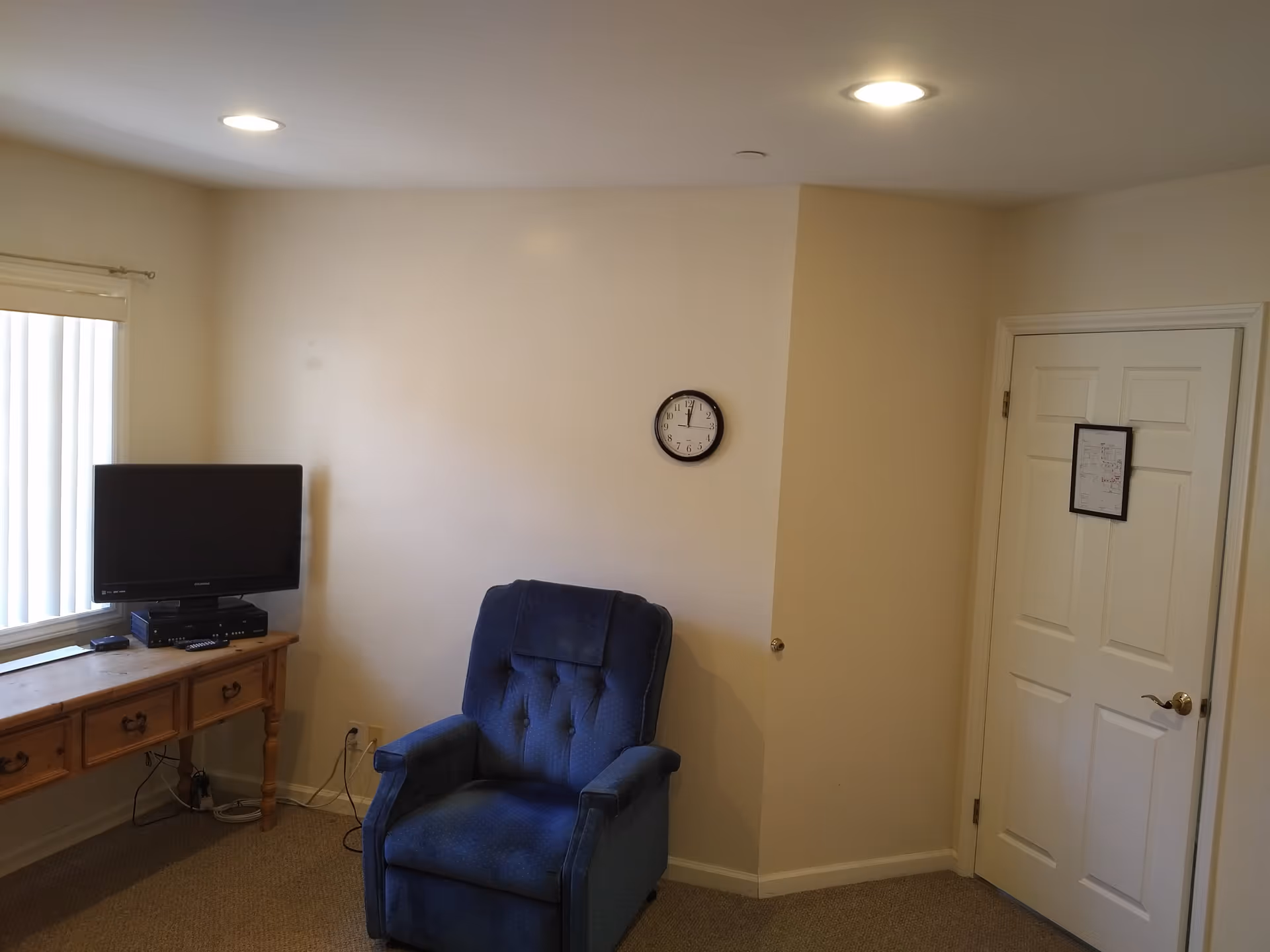 Small living area with a blue upholstered recliner, wall clock, TV on a wooden desk, and a closed door.