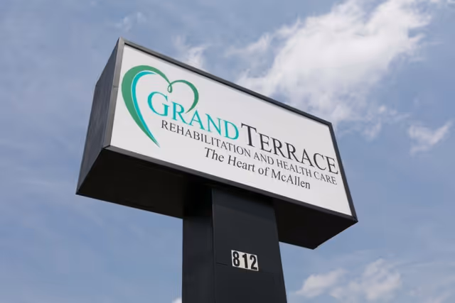 Outdoor sign for Grand Terrace Rehabilitation and Health Care with the tagline 'The Heart of McAllen' against a partly cloudy sky.
