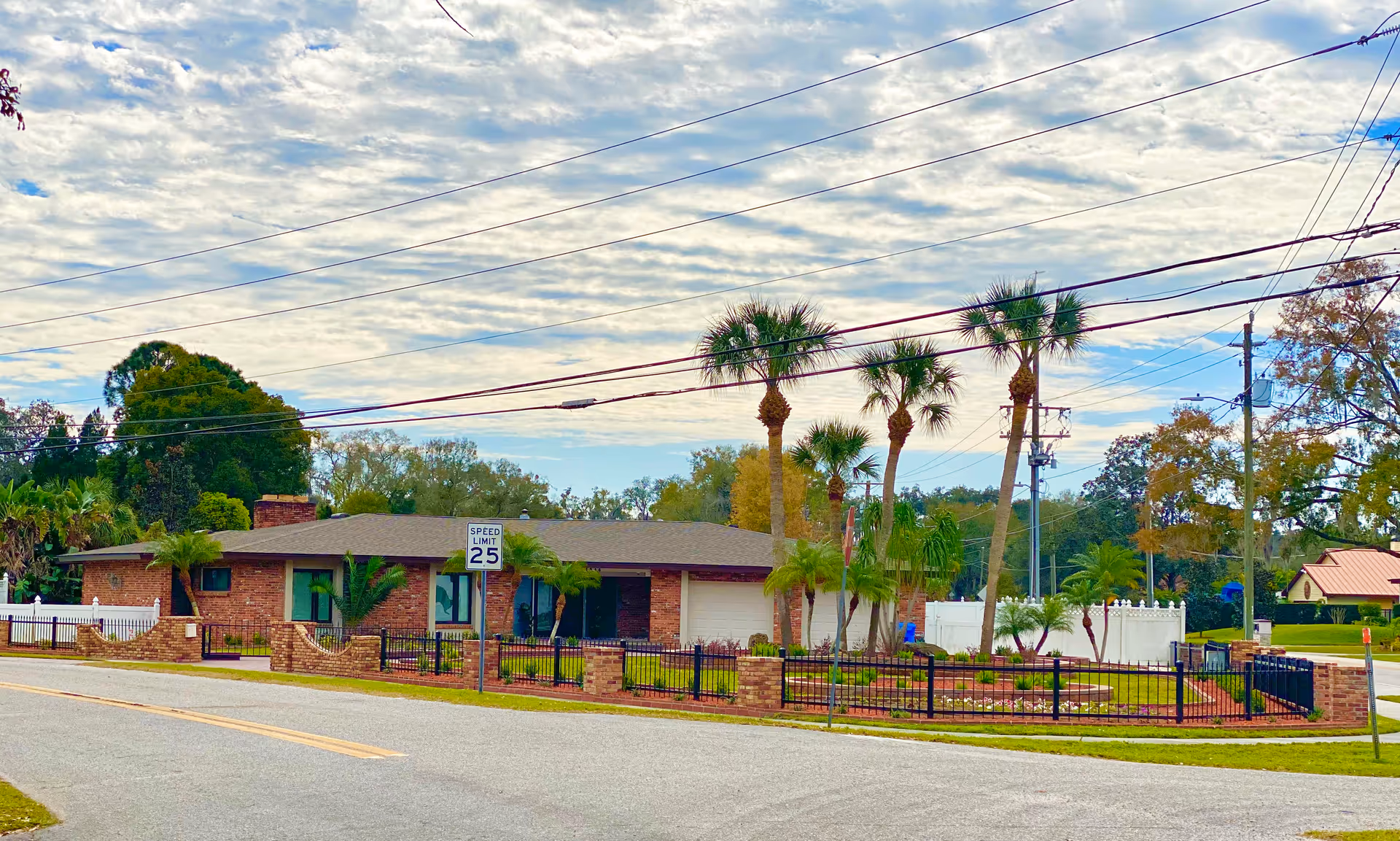 Single-story brick building with palm trees and a fenced front yard along a street with a 25 mph speed limit sign.