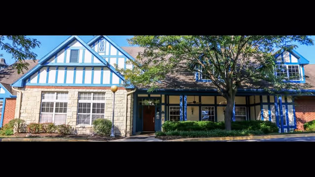Front entrance of a Tudor-style building with blue trim, large windows, a covered porch, and landscaped shrubs under a tree.
