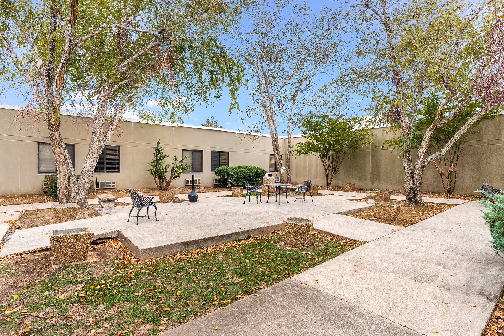 Outdoor courtyard with tables, chairs, trees, and planters beside a beige building.