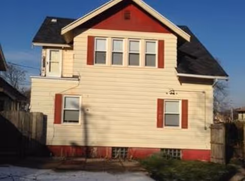 A two-story beige house with red shutters and a fenced yard.