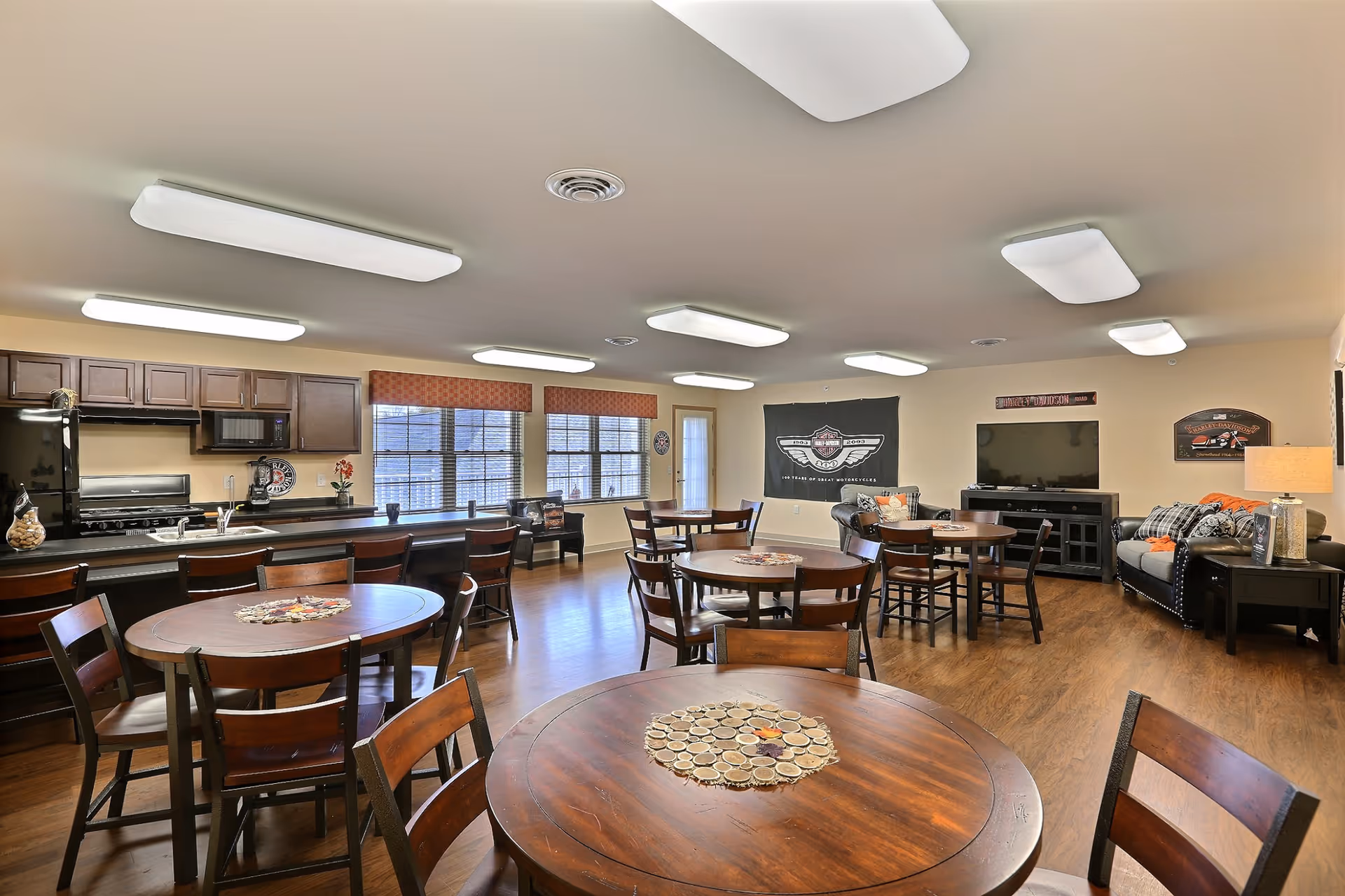 Open communal dining and living area with round wooden tables, a kitchenette along one wall, and seating with a TV and sofa.