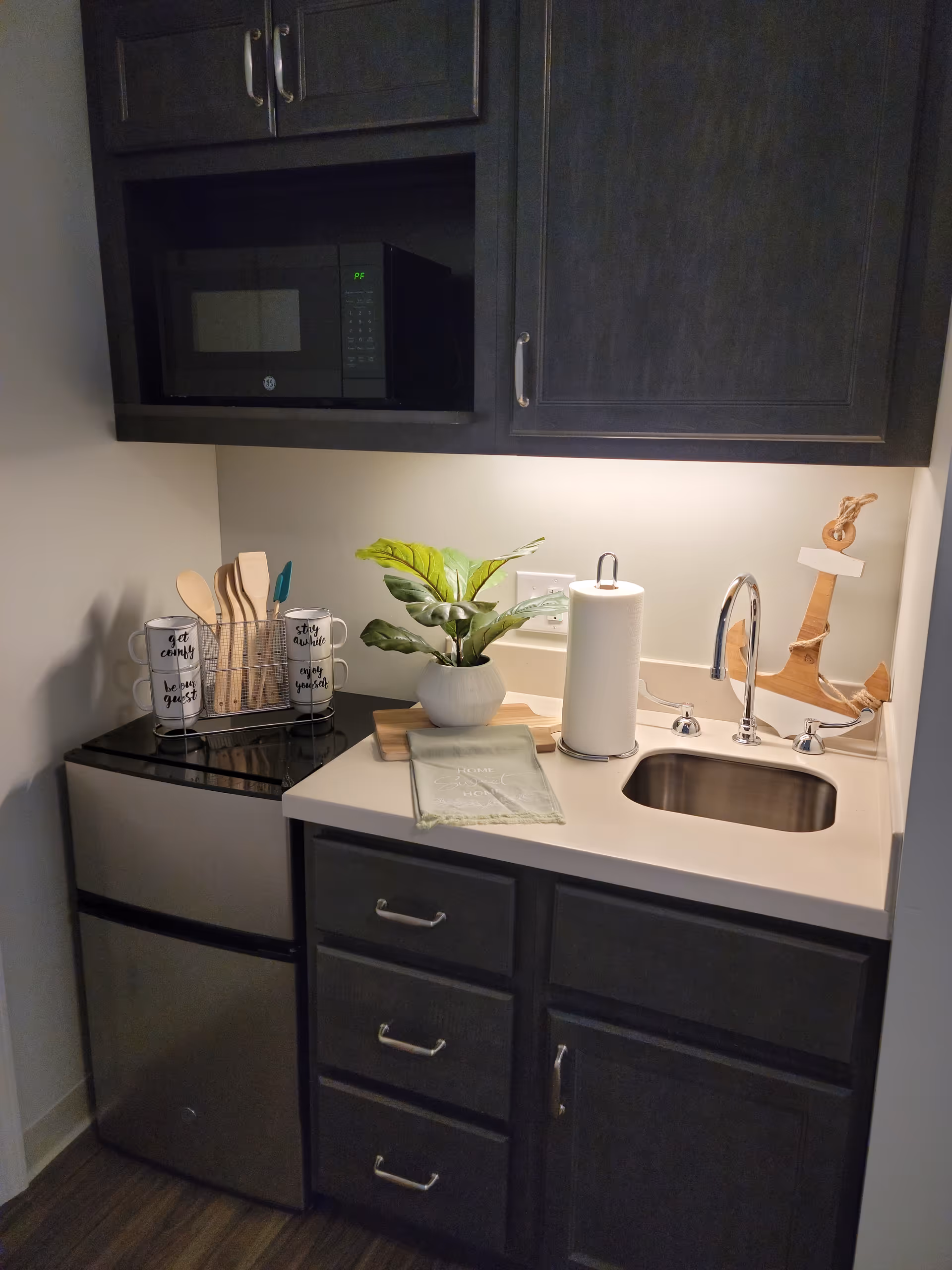 Compact kitchen area with dark cabinets, microwave, sink, plant, and utensils.