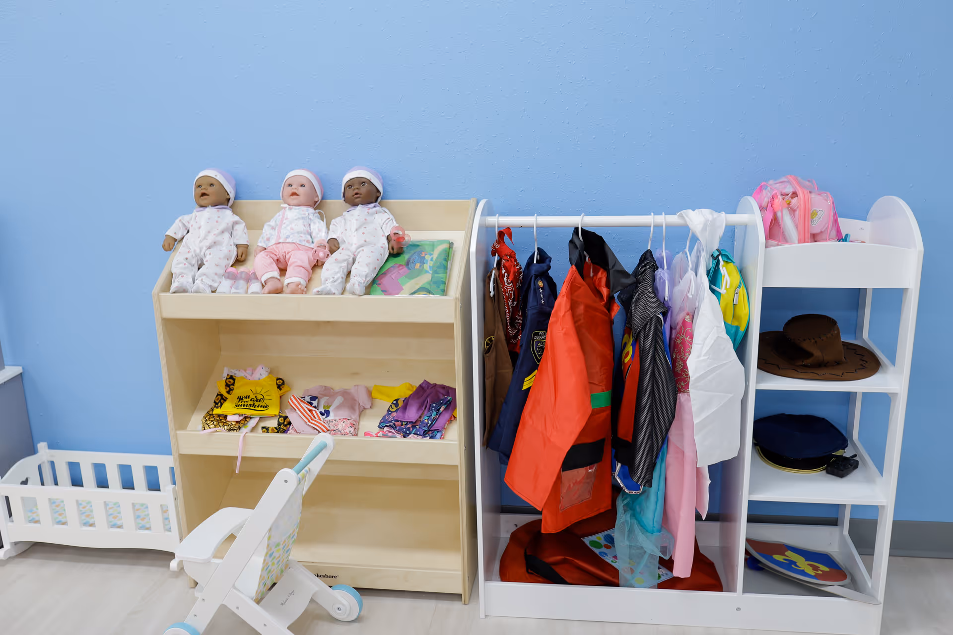 Three baby dolls sit on a shelf beside coat racks in a bright playroom