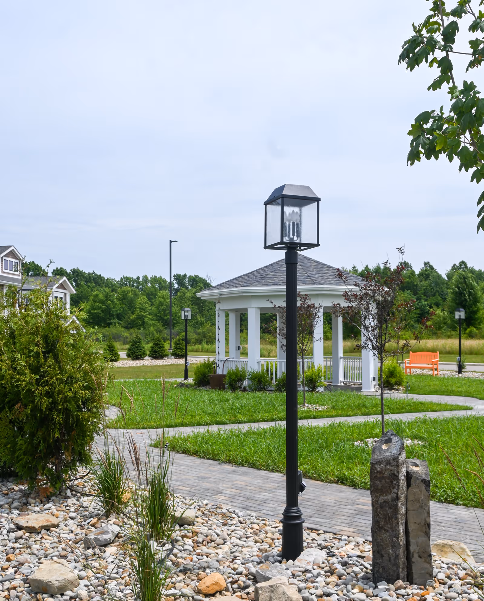 Outdoor garden area with a paved walkway, a black lamp post, a white gazebo with columns, green grass, small trees, bushes, rocks, and an orange bench in the background under a cloudy sky.