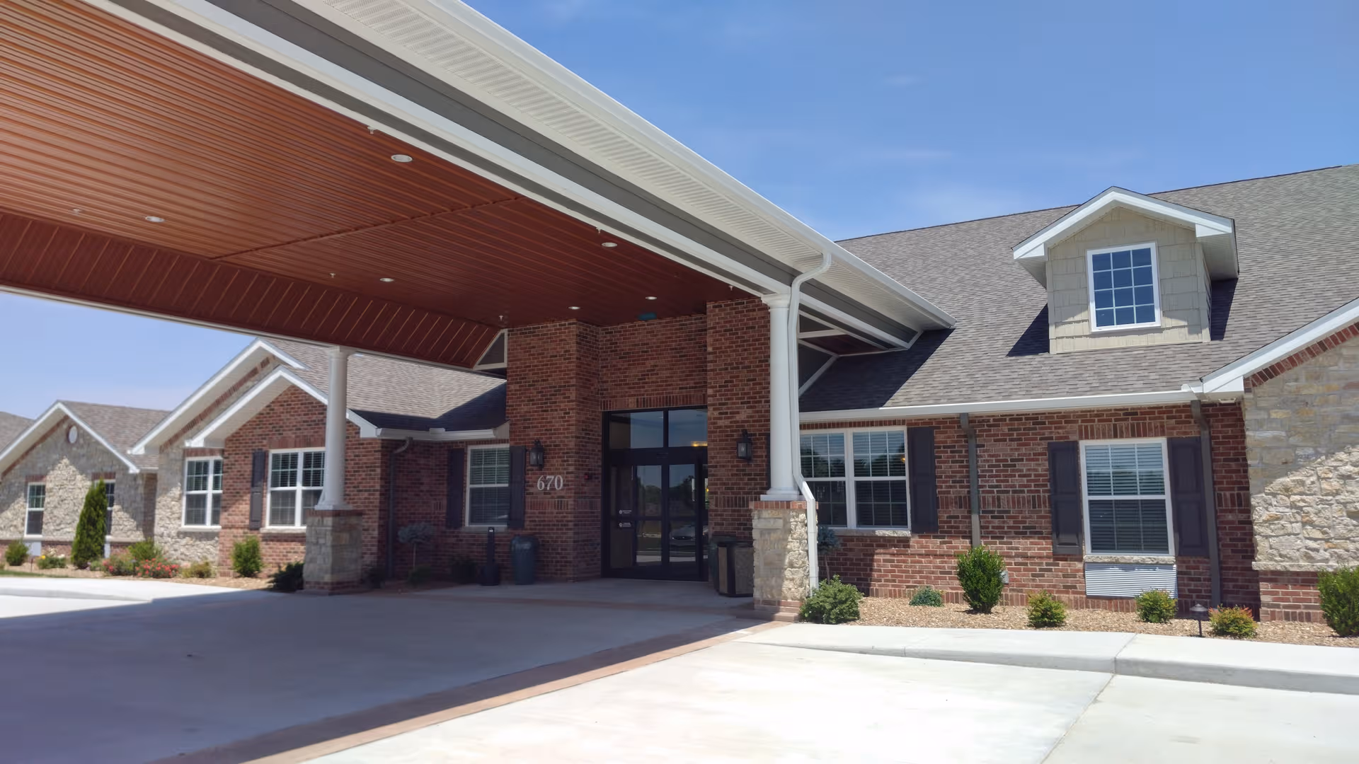 Brick senior living facility with a covered entrance, glass doors, and manicured landscaping.