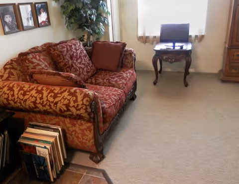 Small living room with a patterned red loveseat, side table under a window holding a laptop, framed photos on the wall and a potted plant.