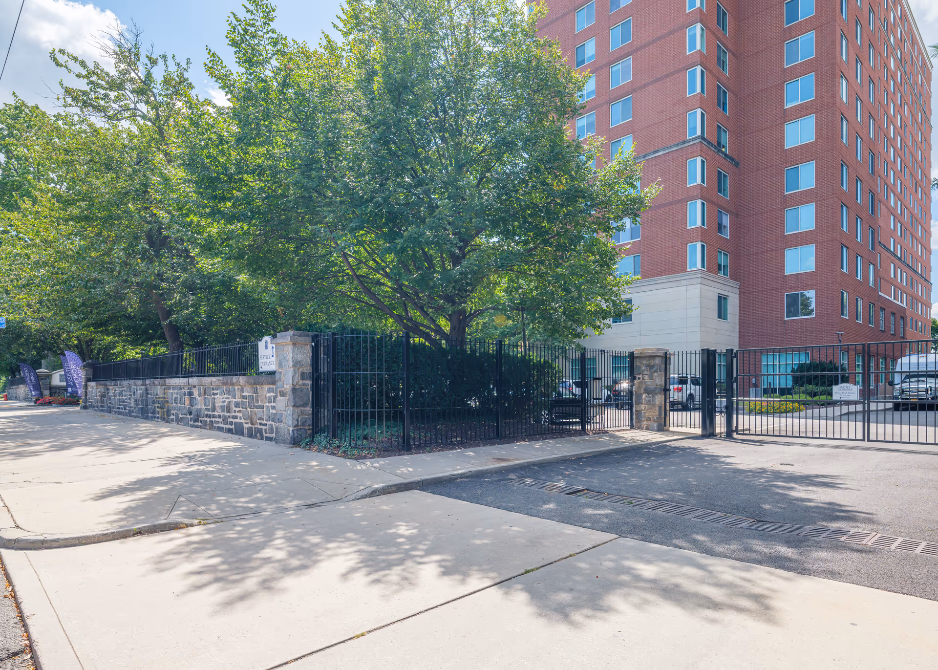 Exterior view of Five Star Premier Residences of Yonkers showing a tall red brick building with many windows, a gated entrance with black metal fencing, stone pillars, and trees providing shade along the sidewalk.