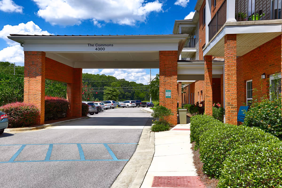 Exterior view of The Commons - 55+ Active Adult Community showing a covered entrance with brick pillars, a parking lot with several cars, and landscaped bushes along the sidewalk under a partly cloudy blue sky.