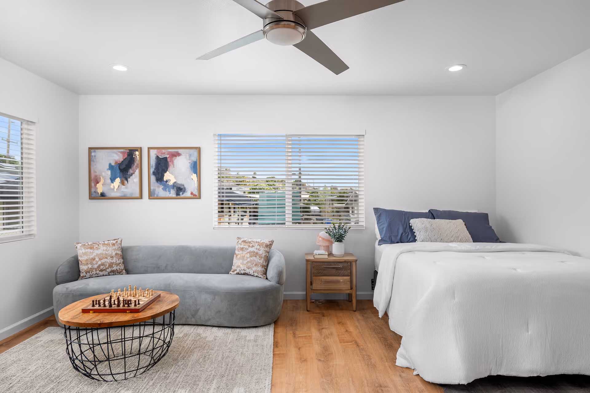 Bright bedroom with a blue sofa, queen bed, window blinds, and wooden side table.