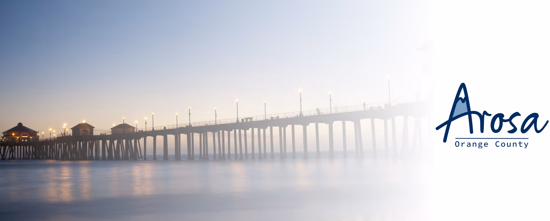 Pier extends over calm water at dusk with lights and Arosa Orange County logo.