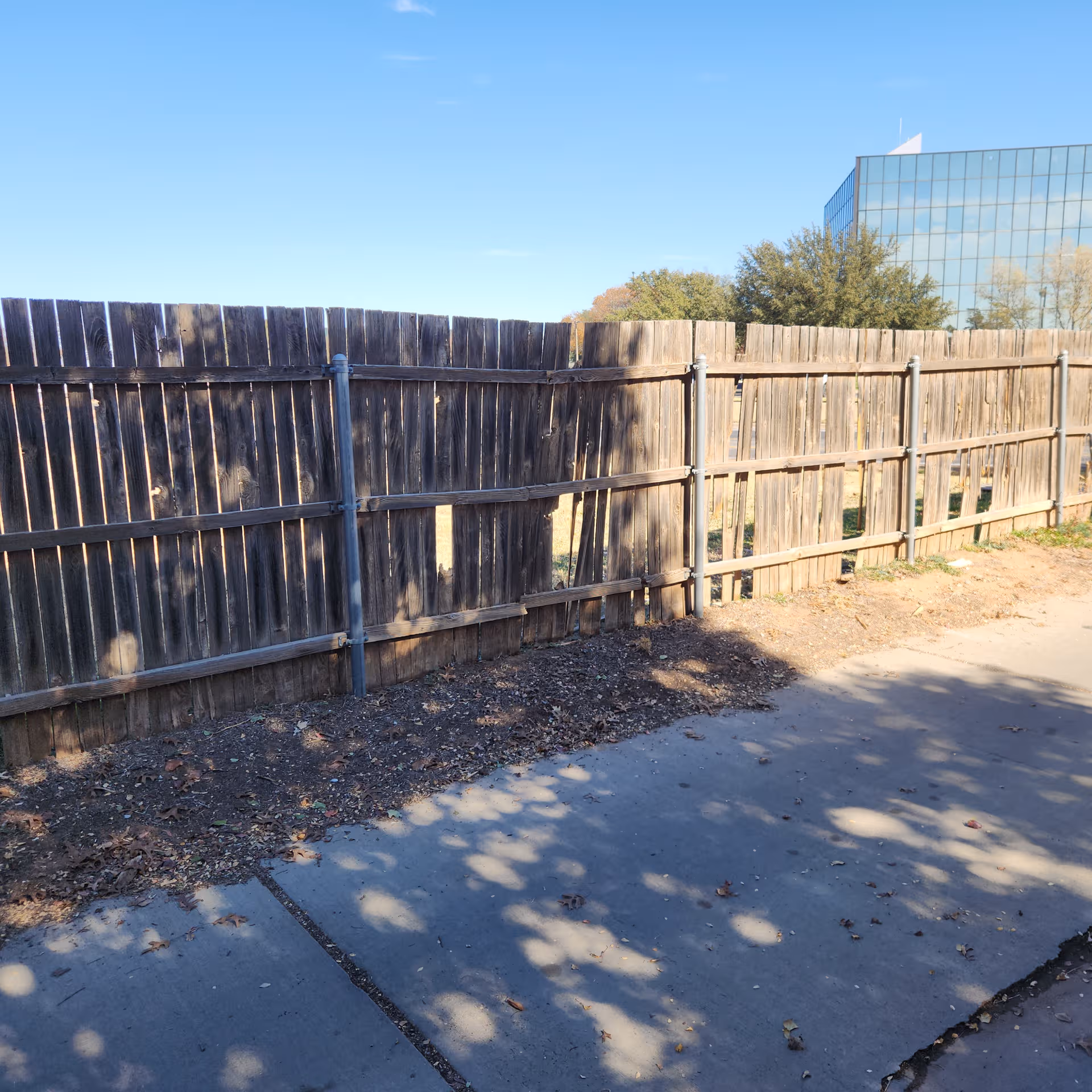 Weathered wooden fence along a concrete sidewalk with a modern glass building in the background.